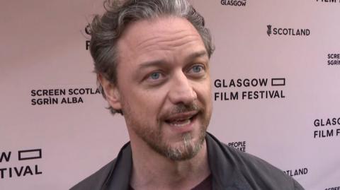 Actor James McAvoy with greying hair and beard, wearing a burgundy jumper and grey jacket. He is standing in front of a Glasgow Film Festival hoarding.