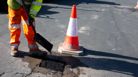 A man in orange reflective trousers and a yellow jacket holding a shovel on a road next to an orange traffic cone and a square of fresh tarmac