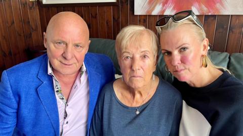 Aldo Zilli, a bald man wearing a blue jacket and light-coloured shirt, sitting next to his mother-in-law Maureen in the centre, and his wife Nichola on the right. They are all sitting on a green leather sofa