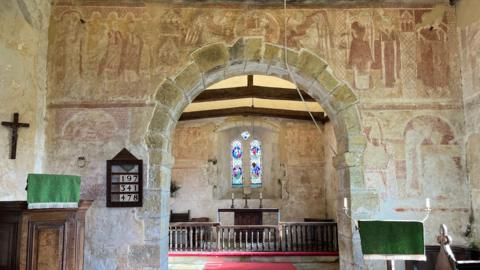 Inside St Botolph's Church which has orange paintings depicting saints and religious scenes on all the walls. The alter and stain glass window can be seen in the centre and the pulpit on the left