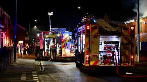 Several fire engines in the street with onlookers standing on a pavement nearby.