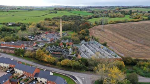Industrial site with stone chimney, set in green fields, shown from the air.