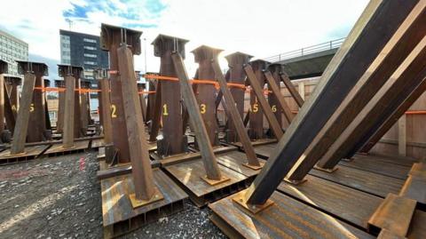 The rust coloured triangular steel props are lined up on ground near to the flyover, which can just be seen in the background. Each prop has yellow letters and numbers on the main, vertical central beam with two thinner props either side of the central section to create the triangle. They are each on top of pallets. In the background are two hi-rise buildings.