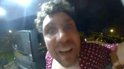 Daniel Lytollis filmed by a doorbell camera. He is wearing a red shirt which is unbuttoned revealing his chest. He has brown messy hair and is staring wide-eyed into the camera.