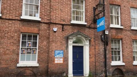 The front of a brick building with large wood-framed windows and a blue door. There is a blue sign hanging beside the door which reads The Hive.