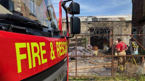 The front of a fire engine is visible on the left, with the words fire and rescue in luminous yellow, with two firefighters and hose reels in the background behind metal fencing at the mill building