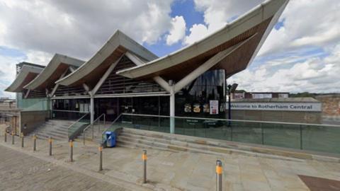 The front of Rotherham Central station, a modern building with sweeping, curved roof sections and large glass walls. Steps and a ramp lead up to the entrance, and a sign on the right reads “Welcome to Rotherham Central”. The area in front is paved with stone and lined with metal bollards.