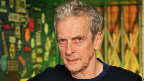 Peter Capaldi has grey hair and is wearing a dark-coloured round-necked top - the wall behind him is multi-coloured 