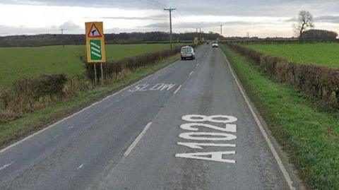 A picture of a road with fields either side. There are two vans in each lane heading in opposite directions.
