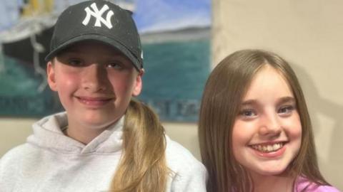 Kendall is wearing a black cap and a grey hoodie. She is on the left of the picture and smiling. Danika is wearing a pink top and has long light brown hair and is smiling on the right of the picture.