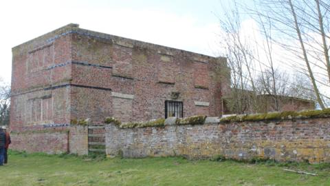 An old brick manor with its windows bricked up, surrounded by gardens and old brick walls.