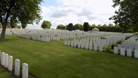 A general view picture of the New Irish Farm cemetery near Ypres. Many rows of white gravestones and lawns are surrounded by trees. 