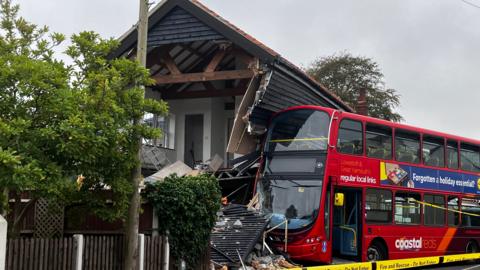 A red double-decker bus has crashed into a house, causing severe damage to the front and side of the property