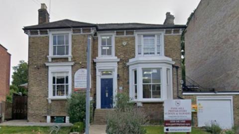 A Google screen grab of a brown brick building with large windows and a blue front door. The signage for the school is in a front garden on the grass.