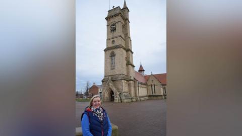 Pamela Hargreaves stands in front of the Christ Church clock tower. It is a stone structure with a clock on it. She has long blonde hair and is smiling at the camera. 