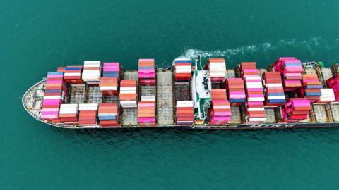 A large cargo ship staked with pink, blue and white container ships sails from right to left through the picture, a small trail of white foam in the picture. 