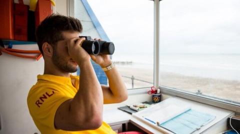 A lifeguard sits in a lifeguard hut on a beach. He looks into binoculars and monitors the beach. He wears a bright yellow t shirt with the letters RNLI written on his arm. He has short brown hair and wears a watch of his left wrist.  