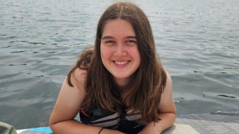 Naomi sat in a lake while paddle boarding. She has long brown hair and is looking directly at the camera and smiling