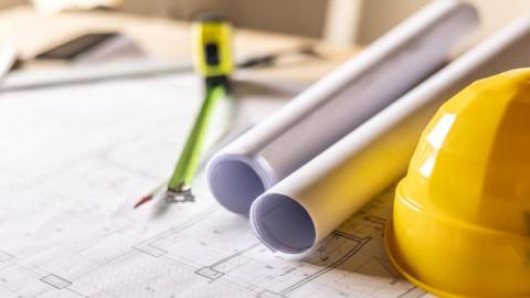 A yellow hard hat sitting on building plants with a neon yellow tape measure.
