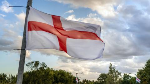 A St George flag, with a red cross on a white background, is flying high up against the sky on a lamppost