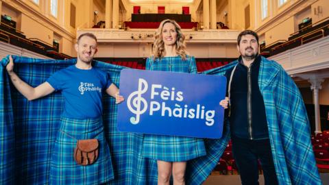 Triùir dhaoine nan seasamh agus plaide thartan timcheall orra.