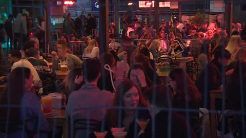 A view of a busy outdoor seating area through a metal fence. Dozens of young people sit drinking, while some of the tables also have shisha pipes. 