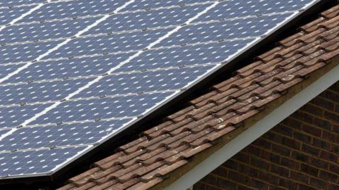 A sloped roof on top of a house which has fitted grid-patterned solar panels on it. It is placed above brown tiles on what looks like a sunny day. 