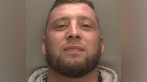 Police mugshot of Adam Shakespeare, who is smiling, wearing a grey jumper with a grey wall in the background