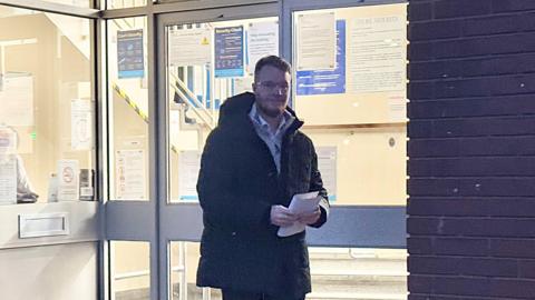 A man with short hair and glasses wearing a thick jacket over a jumper and shirt walking out of court