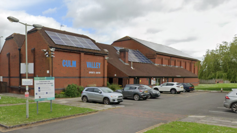 The outside of Culm Valley Sports Centre. It is a red brick building with the words Culm Valley written in large blue writing on one side. The roof is sloped and has solar panels. In front of the building is a car park in which several cars are parked.