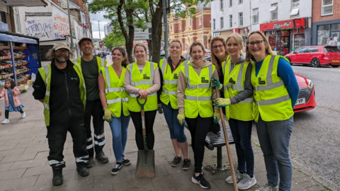 Image shows nine volunteers on Sidwell Street in Exeter. The group is made up of two men and seven women. They are all stood in a line and wearing neon his-vis jackets. The group are smiling and looking at the camera. Two are carrying shovels. 