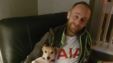 The picture shows a man with short brown receding hair and wearing an open zip hoodie sitting on a sofa with his dog.