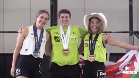 Jules King stand with her arms out holding a Welsh flag grinning with a cowboy hat on. Standing next to her is gold and silver medalists. 