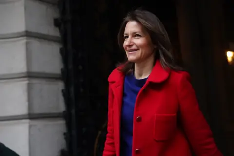 Lucy Frazer in a red coat and blue top walking away from a building 
