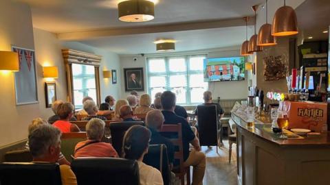 A group of people are sitting indoors in a pub, looking at a screen in the corner of the room. There is a bar to the right hand side of the image. The group is viewed from behind, with people looking towards the screen at the front of the photo.