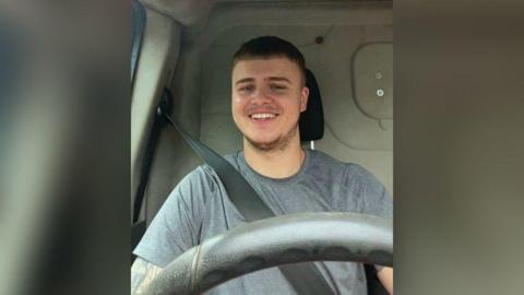 A man is sitting in the driver's seat of a van with his seatbelt on and you can see the steering wheel. He has short brown hair and is wearing a grey top and giving a teethy smile to the camera.