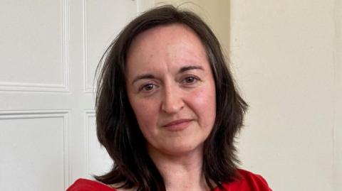 A woman with long, dark hair wearing a red top and sitting in a room with a white panelled wall behind her