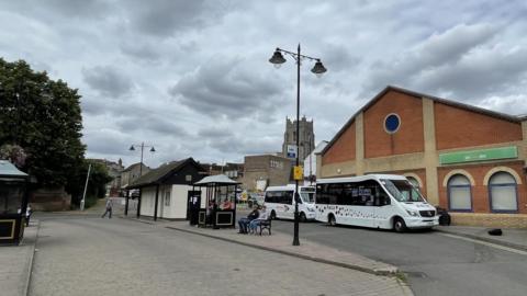 A bus station with people sitting on benches by a road, there is a small pavilion style covering around one bench . Two mini buses are parked behind next to a large brick building.