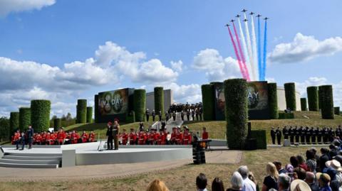 The Red Arrows fly over a topiary garden with a military band performing for onlooking crowds. The jets have red, white and blue streaming behind them through a sunny sky. Banners with VJ DAY 80 are hung from the tall topiary cylinders and representatives of the armed forces in dress uniform stand to attention. 