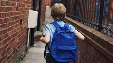 The back of an anonymous child as he walks along a path. He is wearing a blue, short-sleeved school shirt and has a blue backpack.