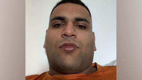 A man in his early 20s wearing an orange T-shirt looking down at the camera