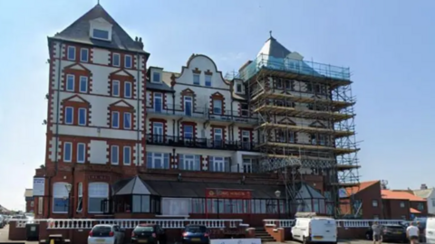 A five-storey Victorian former seafront hotel building with modern bar and conservatory on the ground floor and four floors above, each eight windows across. Three cars and two white vans are parked in parking spaces in front of the building.