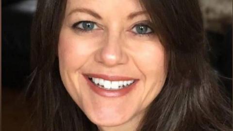 A head shot of Amanda who is looking at the camera and smiling, showing her top front teeth. She has blue eyes and dark brown shoulder length brown hair. The background is blurred and dark