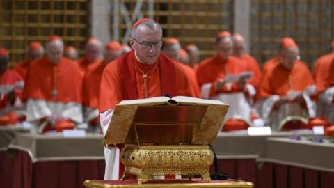 A man in Catholic garb reads from a big book on a lectern while surrounded by other similarly dressed men.