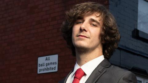 Brad Kella, with mid-length curly brown hair, wears a dark suit with a red tie as he stands in a Liverpool street of terraced houses with a sign on the wall reading "All ball games prohibited"