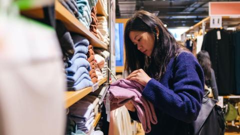 A woman with loose long dark hair in a dark blue jumper is looking at clothes she has taken from a shop shelf