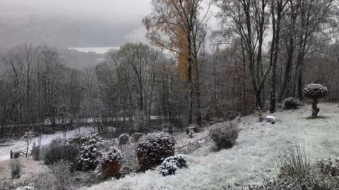 Snow-covered garden and trees on a hillside, with a misty lake and forest in the background under an overcast sky.