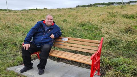 Wayne is wearing a blue coat and sitting on a red bench. The sides are made from a metal design and the slats creating the bench are pale wood.