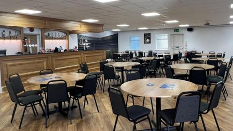 Tables and chairs in an area with a bar and optics behind the bar. The floor in the room is wood and matches the table tops. The room is bright and airy and clean-looking.