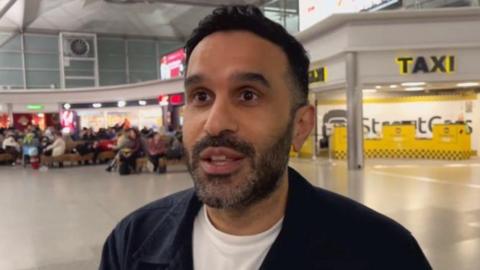 A male passenger with a white shirt and blue jacket looks adjacent to the camera with people sitting down next to a taxi area in Stansted Airport